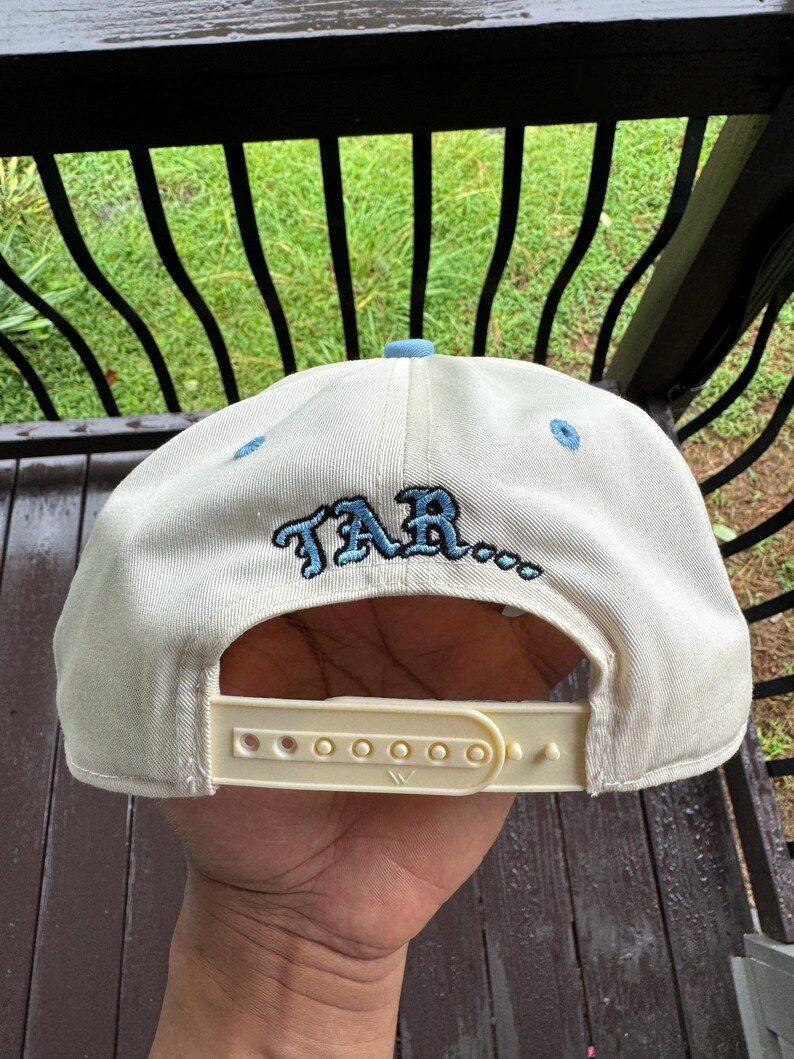 May include: Cream-colored baseball cap with a light blue brim and embroidered text "TAR..." in blue. The cap has a white adjustable snap closure. The cap is made of a durable fabric.