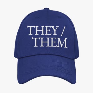 May include: A navy blue baseball cap featuring the white text "THEY / THEM". The cap has a curved peak and a structured crown. The hat is a solid colour and appears to be made of a durable fabric.