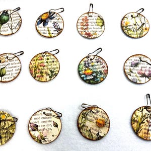 May include: A set of twelve circular tags, each featuring a unique botanical illustration overlaid on vintage French text. Every tag is equipped with a dark metal safety pin attached to the top edge, providing a functional way to secure the paper discs.