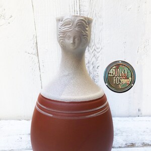 May include: A terracotta vase with a white glaze and a sculpted female head at the top. The vase has a rounded body and a narrow neck.