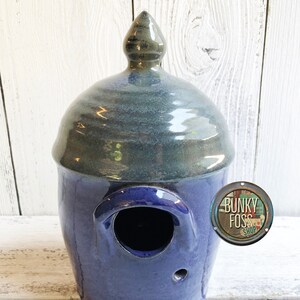 May include: A blue and gray ceramic birdhouse with a rounded top and a small hole for birds to enter. The birdhouse is glazed and has a shiny finish.