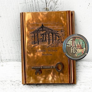 May include: A copper-colored wall hanging with a black and white image of a bridge and the text "Gamble bybro Trondheim". A key is attached to the bottom of the wall hanging.