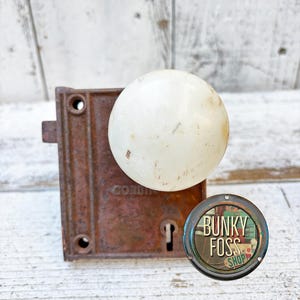May include: A vintage door knob with a white porcelain knob and a rusty metal base. The knob is attached to a metal plate with a keyhole. The plate has the word "CORD" visible on it. The image also includes a round sticker with the text "BUNKY FOSS SHOP".