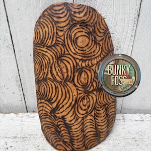 May include: A wooden, oval-shaped cutting board with a carved spiral design. The board is a warm brown color with dark brown carved lines. A circular shop logo is visible on the right side of the board.