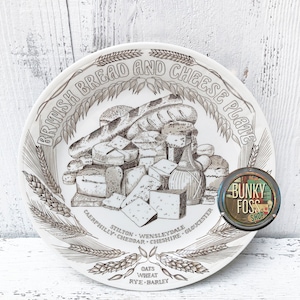 May include: A white ceramic plate with a brown and black illustration of bread and cheese. The plate has the text "British Bread and Cheese Plate" around the top edge and the text "Caerphilly, Stilton, Wensleydale, Cheddar, Cheshire, Gloucester" around the bottom edge. The plate also has the text "Oats, Wheat, Rye, Barley" at the bottom center.