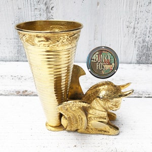 May include: A gold metal vase with a decorative design sits on a white wooden surface. The vase is shaped like a cone and is supported by a gold metal sculpture of a winged creature with horns.