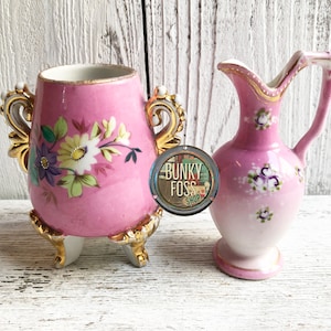 May include: Two pink porcelain vases with floral designs. The larger vase has gold accents and three gold feet. The smaller vase has a handle and a gold rim. Both vases have a white background with pink and purple flowers.