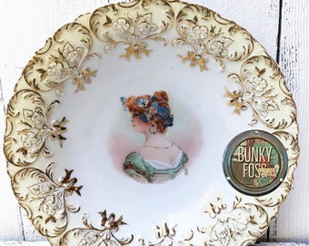 May include: A white porcelain plate with a raised gold floral design around the edge. The center of the plate features a painted image of a woman with red hair wearing a green dress. The plate is in excellent condition.