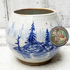 May include: A white ceramic pot with a blue and white landscape design. The pot has a rounded shape and a slightly flared rim. The design features a small island with trees and a body of water. The pot is sitting on a white wooden surface. The text "Bunky Foss Shop" is visible on a metal tag attached to the pot.