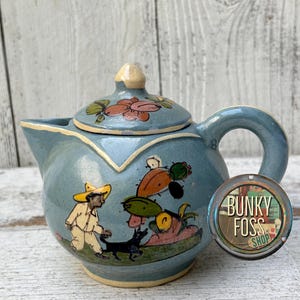 May include: A light blue ceramic teapot with a matching lid, featuring a hand-painted scene of a person in a yellow hat walking a black dog near cacti. The teapot has a circular tag attached that reads BUNKY FOSS SHOP in black and green text.