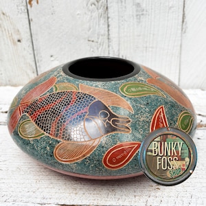 May include: A decorative, oval-shaped vase with a teal background and a black interior. The vase features a detailed fish design in red, black, and silver. Red and green leaf-like shapes are also present. The vase has a pink base.