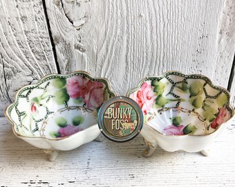 May include: Two small white porcelain bowls with green and pink floral designs and gold accents. The bowls have three small feet and are on a white wooden surface.