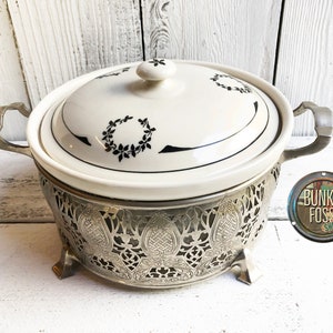 May include: A white ceramic serving dish with a black floral design and a silver metal base with a decorative cut-out pattern. The dish has two handles and a lid.