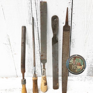 May include: Five vintage metal files with wooden handles. The files are different sizes and have various patterns on the metal surface. The files are arranged on a white wooden surface.