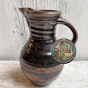 May include: A brown ceramic pitcher with a wide mouth and a handle. The pitcher has a rustic, hand-thrown look with a dark brown glaze and a hint of orange.