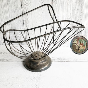 May include: Vintage metal wire basket with a handle. The basket has a rounded base and a flared, open design. The metal has a dark, aged finish. The basket is likely used for displaying fruit or other items. The basket is on a white wooden surface.