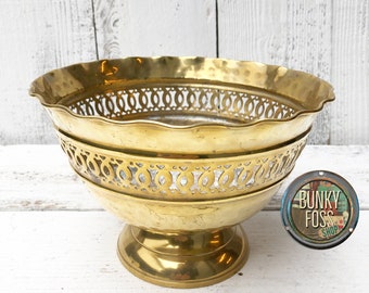 May include: A gold metal bowl with a decorative, openwork design around the rim and sides. The bowl has a scalloped edge and sits on a pedestal base.