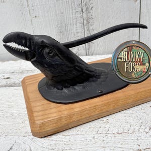 May include: A black cast iron nutcracker shaped like a bird's head with a wooden base. The nutcracker is on a wooden base with a sticker that says "Bunky Foss Shop".