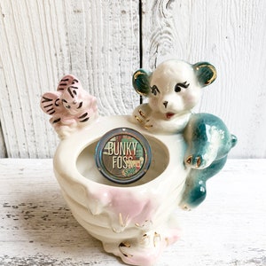May include: A vintage ceramic planter shaped like a stack of plates with a bear and a bird figurine on top. The planter is white with pink and gold accents. The bear is blue and white with gold accents. The bird is pink and white with black accents. The planter has a small hole in the center for drainage. The planter is decorated with the words "Bunky Foss Shop".