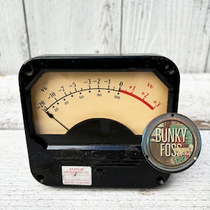 May include: A black vintage VU meter with a white face and red needle. The meter is labeled "VU" and has markings from -20 to +3. The meter is made by Branom Instrument Co. and is from Seattle, Portland. A round metal button with the text "Bunky Foss Shop" is attached to the meter.