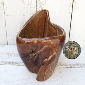 May include: A brown ceramic planter with a unique, abstract shape. The planter has a pine cone design and a glossy finish.