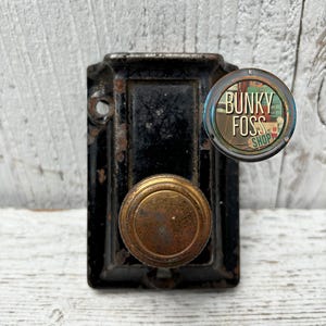 May include: Black metal door knob with a brass handle and a round, black metal plate. The plate has two small holes. The knob is attached to a white wooden surface.