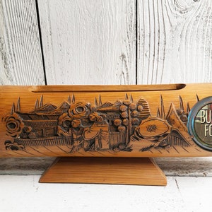 May include: A carved wooden planter in the shape of a boat with a scene of a village and people. The planter is made of bamboo and has a natural wood finish. It is sitting on a wooden stand.