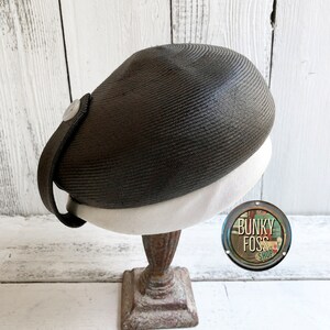 May include: A black and white straw hat with a white band and a button on the side. The hat is on a rusty metal stand. The hat is labeled "Bunkie Foss Shop".