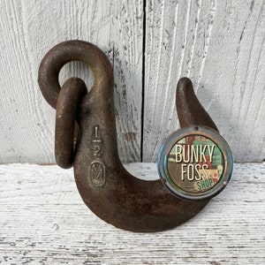 May include: A rusty metal hook with a large ring at the top. The hook is marked with the number "1 2" and a letter "M". A round sticker with the text "Bunky Foss Shop" is attached to the hook.