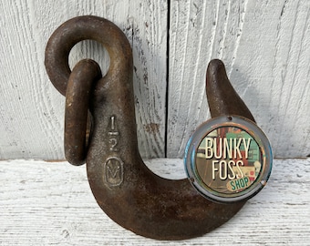 May include: A rusty metal hook with a large ring at the top. The hook is marked with the number "1 2" and a letter "M". A round sticker with the text "Bunky Foss Shop" is attached to the hook.