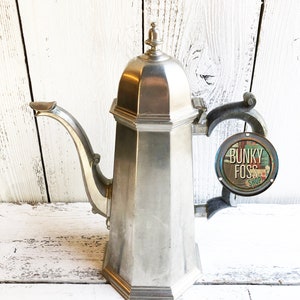 May include: A vintage silver-toned metal coffee pot with a unique geometric design. The pot features a long, curved spout, a hinged lid, and a decorative handle. A small sign that says "Bunky Foss Shop" is attached to the handle.