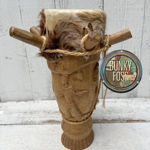 May include: A wooden drum with a carved design and a fur-covered top. The drum has two wooden handles and is tied with rope. The drum is labeled "Bunky Foss Shop".