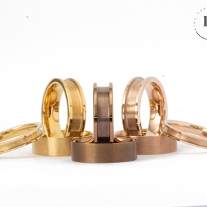 May include: A set of six wedding bands in different colours, including gold, rose gold, and a darker metal. The bands are all polished and have a bevelled edge.