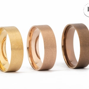 May include: Three brushed metal wedding bands in gold, rose gold, and brown. The bands are all the same width and have a matte finish.