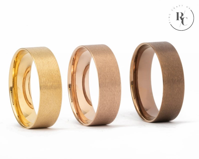 May include: Three brushed metal wedding bands in gold, rose gold, and brown. The bands are all the same width and have a matte finish.