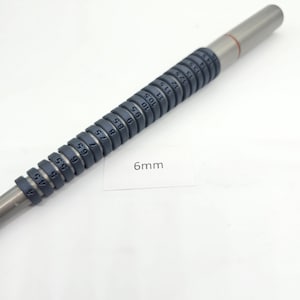 Puede incluir: Una herramienta de medición de anillos de metal plateado con una serie de calibradores de anillos de color gris oscuro. Cada calibre está etiquetado con un número de tamaño. El texto "6mm" está en una etiqueta blanca. Herramienta para medir tallas de anillos.