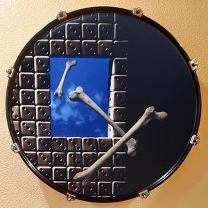 May include: A round drumhead with a surreal design. The image features a square window revealing a blue sky with bones floating in it. The left side has a pattern of dice, while the right side is black.