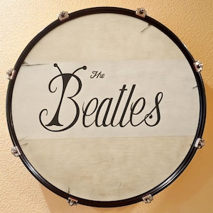 May include: A round drumhead with a black rim and silver hardware. The drumhead features the words "The Beatles" in a stylized black font on a white background. The drum is likely a decorative item or a piece of music memorabilia.