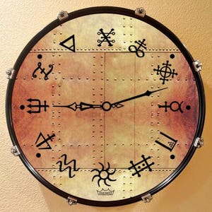 May include: A round wall clock with a steampunk design. The clock face has a metallic, riveted appearance with black symbols marking the hours. Black clock hands point to the time. The clock is framed in black with silver hardware.