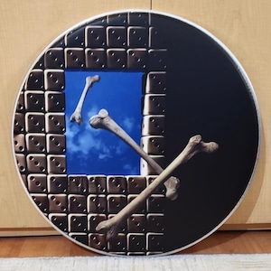 May include: Round wall art featuring a surreal design. The artwork depicts a window-like frame with a dice pattern, revealing a blue sky with floating bones. The right side is black, creating a contrast. The overall theme is abstract and thought-provoking.