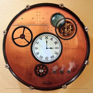 May include: A steampunk-style wall clock with a copper-colored face and black trim. The clock features gears, a round dial with unique symbols, and the words "time machine". The clock is a unique decorative item.