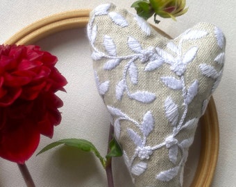 May include: A white heart-shaped cushion with white embroidery of leaves and vines on a beige linen fabric. The cushion is displayed on a wooden embroidery hoop.