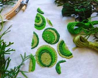 May include: A close-up of a white linen fabric with a green embroidered design of a stylized Christmas tree. The tree is made up of circles and semi-circles, and there are spools of green embroidery floss nearby.
