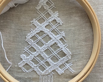 May include: A white embroidery hoop with a white thread and a white cross-stitch design of a Christmas tree on a beige linen fabric.