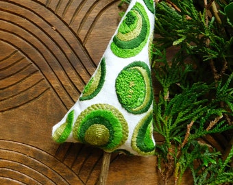 May include: A white fabric Christmas tree ornament with green embroidered circles. The ornament is on a wooden surface with a green leafy branch.