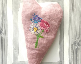 May include: A pink heart-shaped decoration with embroidered flowers. The embroidery shows a daisy, a rose, and small blue flowers with green stems. The heart is set against a watercolour background and framed in a grey wooden frame.