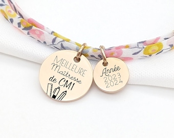 Puede incluir: Un brazalete de oro con una banda de tela con estampado floral y dos dijes. Un dije dice "Meilleure Maitresse de CM1" y el otro dice "Annee 2023-2024".