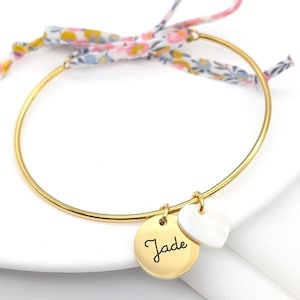 Peut inclure: Un bracelet jonc en or avec un charme qui dit "Jade" et un charme en forme de cœur blanc. Le bracelet est attaché avec un ruban floral.