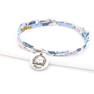 Könnte beinhalten: Ein Stoffarmband mit hellblauem Blumenmuster und einem runden, silberfarbenen Metallanhänger. Der Anhänger ist mit einer Illustration eines offenen Buches, umgeben von einem Blumenkranz, und dem Wort Merci in Schreibschrift graviert.