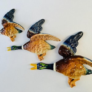 May include: Three ceramic duck wall decorations in flight. The ducks have brown, tan, and blue plumage with yellow beaks and green heads. The wings are outstretched, and the ducks appear to be in motion.